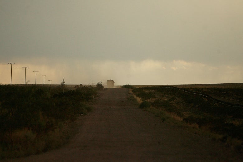 A bus is seen in the distance on a dirt road through the dust and sparse brush of a desert at dusk.
